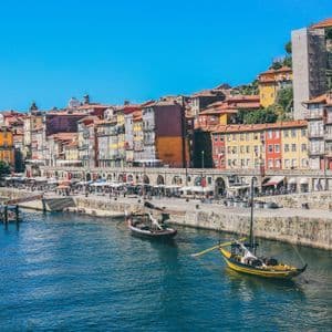 Barche tradizionali galleggiano su un fiume accanto a un lungomare in pietra, con edifici colorati a più piani, sotto un cielo azzurro e limpido.