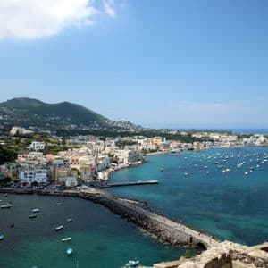 Una colorata cittadina costiera con un ponte di pietra, vista dall'alto, con molte barche nell'acqua blu e una collina verde sullo sfondo.