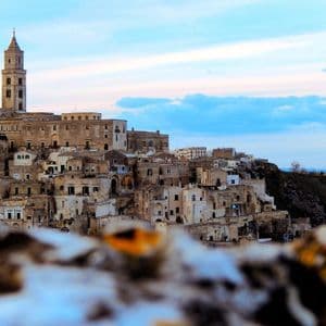 Un borgo in pietra con un campanile imponente, arroccato su una collina, visibile oltre un primo piano sfocato e sotto un cielo dai colori pastello.