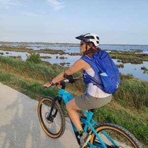 Una donna con casco e zaino percorre un sentiero in bicicletta blu acceso, accanto a una grande palude, sotto un cielo sereno.