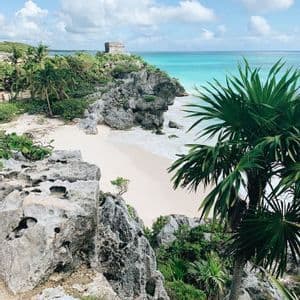 Une ancienne ruine de pierre sur une falaise rocheuse surplombe une plage de sable blanc et une mer turquoise, avec des palmiers au premier plan.