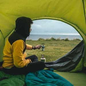Eine Person in einem gelb-schwarzen Kapuzenpullover kocht Kaffee mit einer Mokkakanne, während sie in einem Zelt mit Meerblick sitzt.