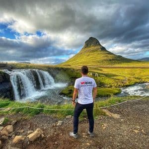 From behind, a WeRoad group leader looks out at a waterfall cascading next to a large, green, cone-shaped mountain.