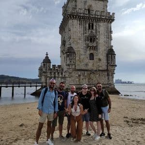 Eine WeRoad-Gruppe von sieben Personen posiert an einem Sandstrand vor einem großen Steinturm am Wasser.