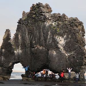 Un groupe WeRoad en voyage prend la pose sous une grande formation rocheuse arquée sur une plage de sable noir.