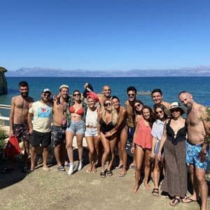Un grand groupe WeRoad en voyage posant pour une photo sur une plage ensoleillée, avec une falaise rocheuse et la mer bleue en arrière-plan.