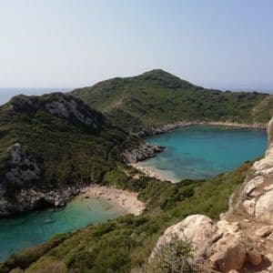 A high-angle view of a twin cove with turquoise water and sandy beaches, nestled between lush green hills under a clear sky.