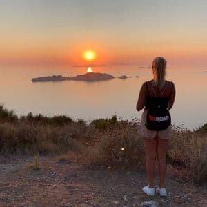 Una donna con uno zaino WeRoad si trova su una collina, osservando il tramonto sul mare e isole lontane.