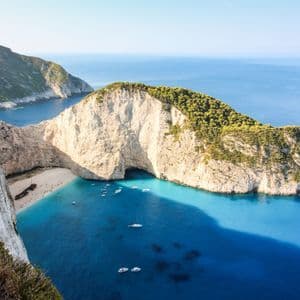 Vista aerea di una caletta appartata con un relitto su una spiaggia di sabbia bianca, circondata da scogliere e acque turchesi con barche.