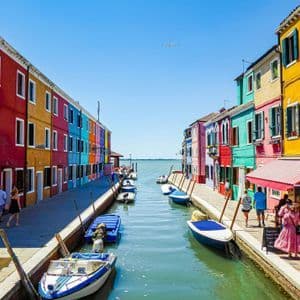 Un canal étroit avec des bateaux amarrés sépare les quais bordés de maisons aux couleurs vives, tandis que des gens se promènent par une journée ensoleillée.