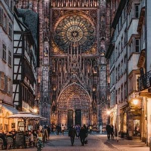 Una veduta di una strada acciottolata al crepuscolo che conduce alla facciata illuminata di una grande e ornata cattedrale gotica con un imponente rosone.