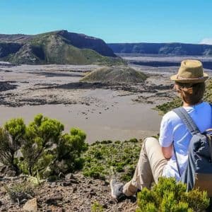 Una persona con uno zaino e un cappello è seduta su un'altura verde che si affaccia su una vasta caldera vulcanica in una giornata di sole.