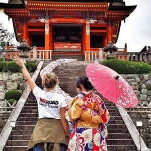 Un capogruppo WeRoad è in piedi con una donna in kimono tradizionale e ombrellino alla base di scale che conducono a un tempio arancione.