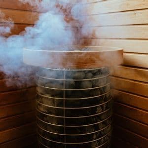 Il vapore sale dalle pietre calde contenute in una stufa per sauna cilindrica in metallo, posta contro una parete rivestita in legno.