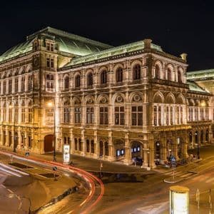 Un bâtiment historique orné, illuminé la nuit au coin d'une rue de ville, avec des traînées lumineuses dues au trafic en pose longue.