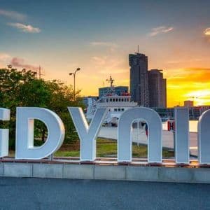 Una grande insegna bianca con la scritta 'GDYNIA' su una piattaforma in riva all'acqua, con lo skyline della città e un tramonto colorato sullo sfondo.