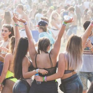 Diverse donne di spalle con le braccia alzate in una folla a un festival all'aperto soleggiato.