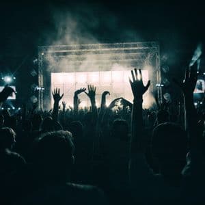 Sagome di una folla con le mani alzate davanti a un palco illuminato a festa durante un concerto notturno.
