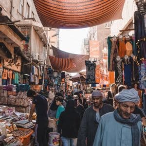 Una multitud de personas recorre un estrecho callejón de mercado soleado, lleno de puestos que venden ropa y artículos bajo un toldo a rayas.