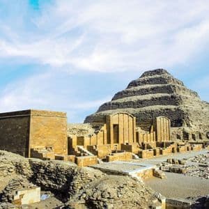 Eine große Stufenpyramide steht zwischen alten Steinruinen in einer Wüstenlandschaft unter einem teils bewölkten blauen Himmel.