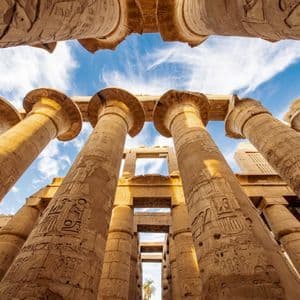 A low-angle view of massive ancient columns with hieroglyphic carvings reaching towards a blue sky with white clouds.