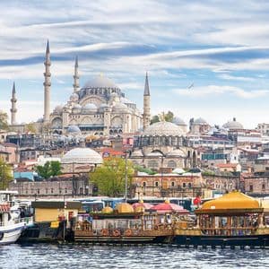 Eine große Moschee mit Kuppeln und Minaretten überblickt eine städtische Uferpromenade, an der mehrere Boote festgemacht sind.