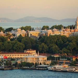 A historic palace complex with domes and towers sits on a lush, green hill overlooking the water at sunset.