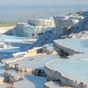 Sunlight illuminates white travertine terraces filled with pools of pale blue water on a hillside.