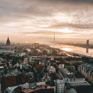 Eine Luftaufnahme einer Stadtsilhouette neben einem breiten Fluss während eines diesigen Sonnenaufgangs, mit verschiedenen Gebäuden und einem hohen Turm in der Ferne.
