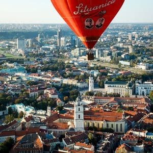 Un pallone aerostatico rosso con la scritta 'Lituanica' sul fianco fluttua sopra un vasto paesaggio urbano con edifici storici dai tetti rossi.