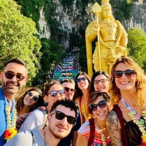 Un viaggio di gruppo WeRoad scatta un selfie davanti a una grande statua dorata e una scala colorata che sale su una scogliera rigogliosa e rocciosa.