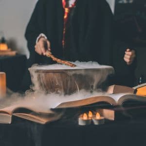 Una persona in tunica nera punta una bacchetta magica su un calderone fumante su un tavolo con libri aperti e candele accese.