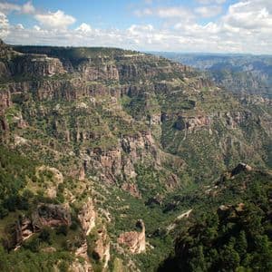 Una vista panorámica de un vasto sistema de cañones con acantilados rocosos y empinados, y laderas cubiertas de árboles y arbustos verdes bajo un cielo azul parcialmente nublado.