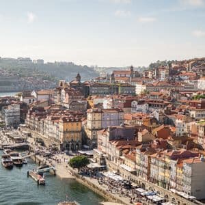 A high-angle view of a city with colorful buildings and terracotta roofs built on a hillside next to a wide river.