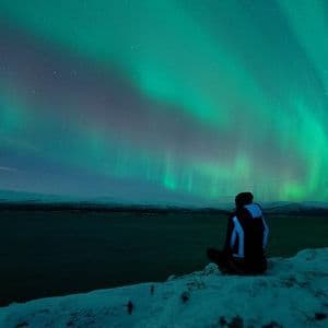 Une personne est assise sur une falaise enneigée, observant les aurores boréales vertes illuminer le ciel nocturne étoilé au-dessus d'une étendue d'eau sombre.