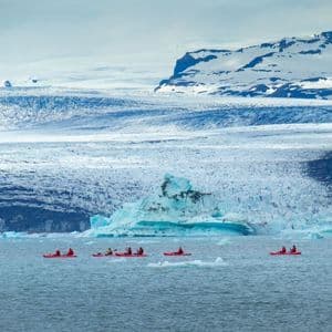A WeRoad group trip paddles in red kayaks on a glacial lake, with a massive glacier and snowy mountains in the background.