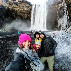 Un viaje en grupo de WeRoad de cuatro personas con ropa de invierno se toma una selfie junto a una cascada en un terreno nevado de arena negra.