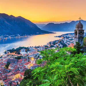 Vista panoramica di una città costiera e della baia al tramonto, con un campanile in pietra in primo piano e montagne sullo sfondo.