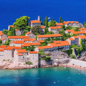 Veduta dall'alto di un villaggio isolano con case in pietra e tetti rossi, delimitato da una spiaggia sabbiosa e dal mare blu.