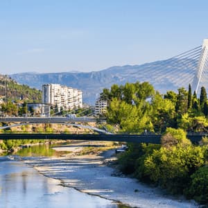 Un moderno ponte strallato e altri ponti più piccoli attraversano un fiume in una città circondata da alberi verdi e montagne distanti.