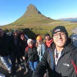 Un groupe WeRoad prend un selfie souriant devant une grande montagne verte et une rivière par une journée ensoleillée.