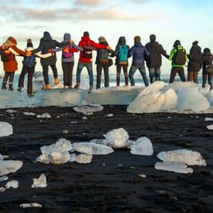 Un gruppo WeRoad in viaggio, a braccetto, è in piedi su grandi blocchi di ghiaccio su una spiaggia di sabbia nera, ammirando il mare.