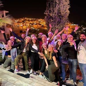Un gruppo WeRoad in viaggio, con kefiah, posa per una foto notturna su una scalinata, con una città illuminata sullo sfondo.