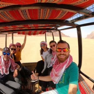 A WeRoad group trip smiles for a selfie in the back of an open-air vehicle during a tour of a desert with rocky mountains.