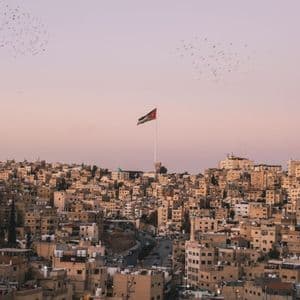 The flag of Jordan flies over a dense cityscape of sand-colored buildings under a pale pink sky with birds flying.