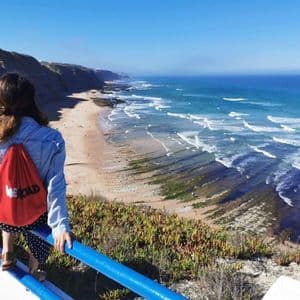 Eine Frau mit einem roten WeRoad-Rucksack steht auf einer Klippe und blickt auf einen langen Sandstrand und die Meereswellen.