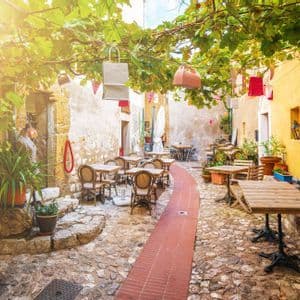 Un caffè all'aperto vuoto in una stretta strada di ciottoli, con tavoli e sedie disposti sotto un pergolato di viti verdi e frondose.