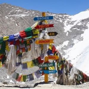 Un cartel adornado con coloridas banderas de oración indica direcciones a lugares como Siachen, Leh y Kargil en un puerto de montaña nevado.