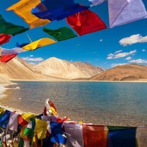 Coloridas banderas de oración cuelgan sobre la orilla de un lago tranquilo, con grandes montañas áridas al fondo bajo un cielo azul.