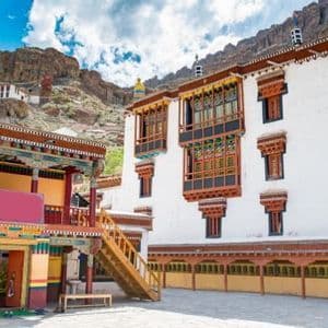 Un monasterio tradicional y ornamentado, con muros blancos y ventanas de madera de colores, se erige en un patio al pie de una montaña rocosa.